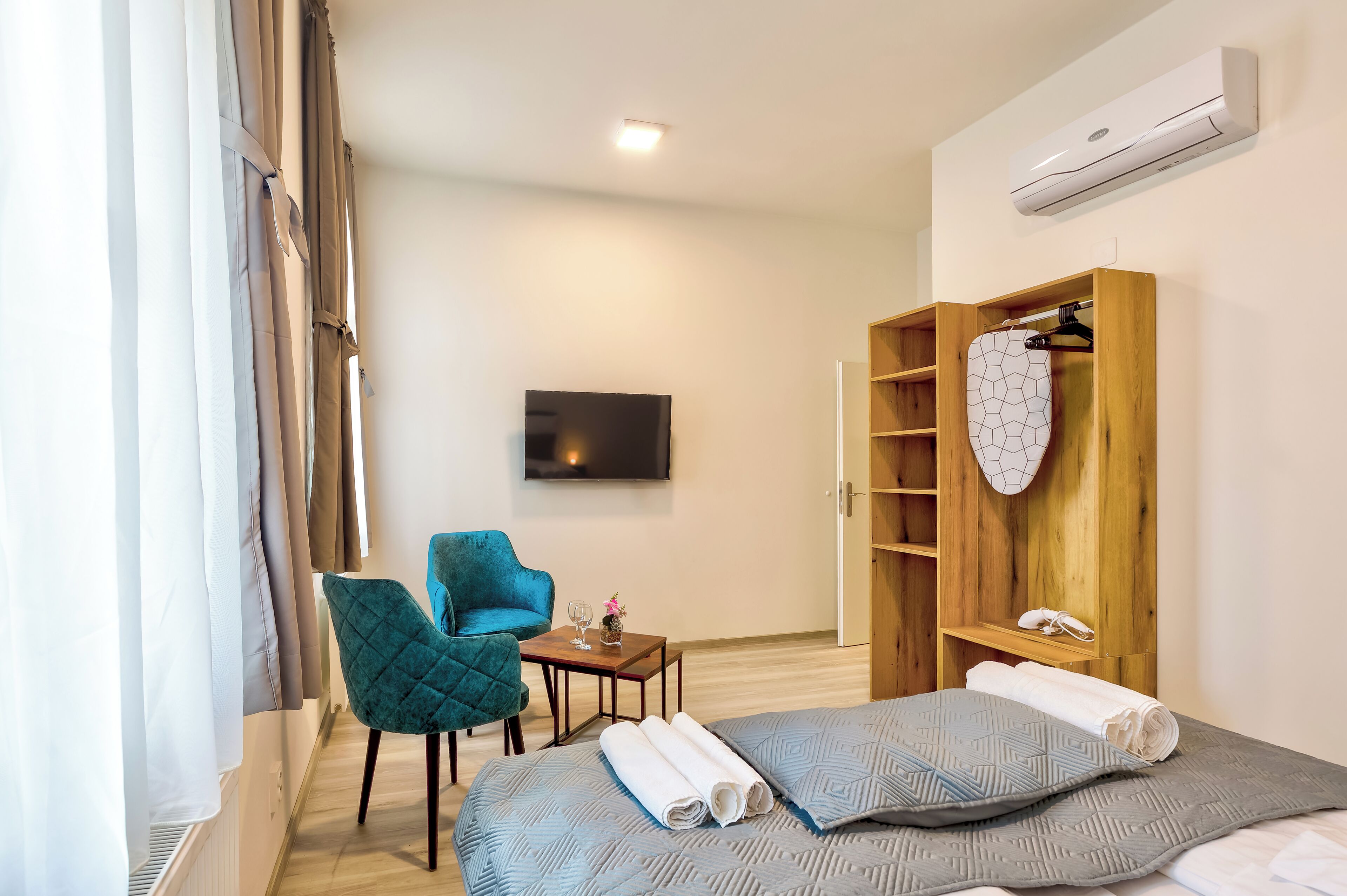 A hotel room with a bed, a blue chair, a small table with a glass, and a wooden wardrobe with a heart-shaped mirror.