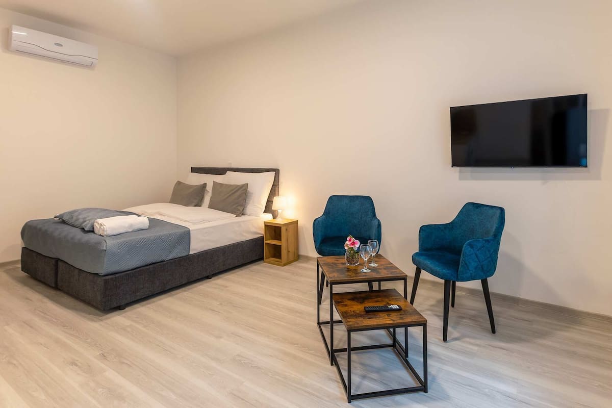 A bedroom with a bed, bedside table, two chairs, a TV, and an air conditioning unit.