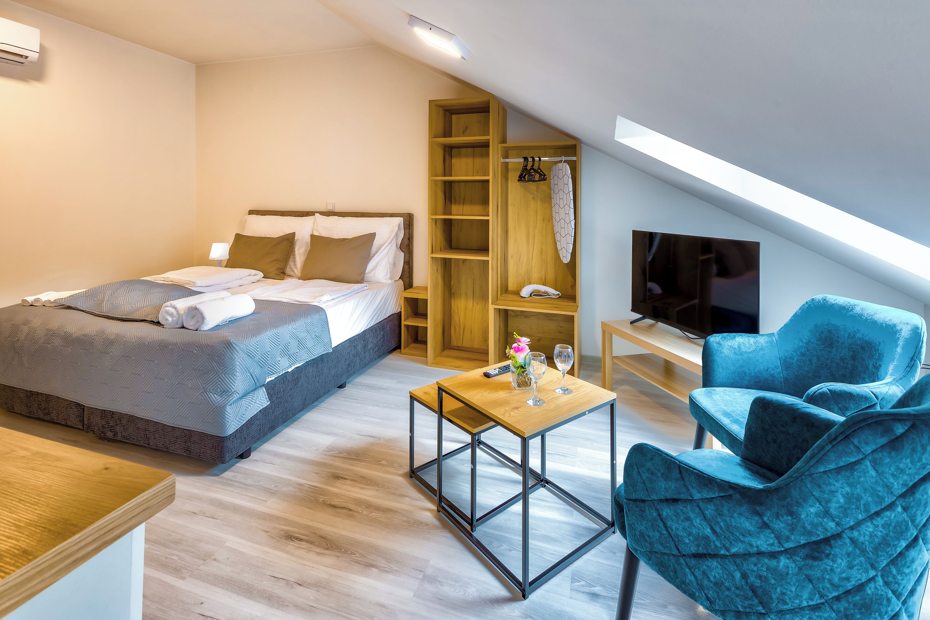 Ein modernes Schlafzimmer mit einem Bett, einem Holzschrank, einem kleinen Tisch mit Fernbedienung, einem blauen Sessel und einem Fernseher auf einem Holzboden.