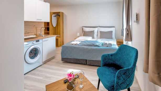 A modern hotel room with a bed, a blue chair, a washing machine, and a wooden table with a vase and a remote control.