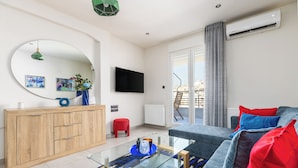 A modern living room with a glass coffee table, a grey sofa with red and blue cushions, a wooden cabinet, a large mirror, and a flat-screen TV. - Aether by halu (Thessaloniki)
