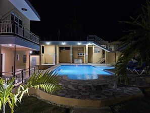 Ein Schwimmbecken mit Fliesen umgeben, ein überdachter Bereich mit einer Bar und eine Treppe, die zu einem Balkon führt.
