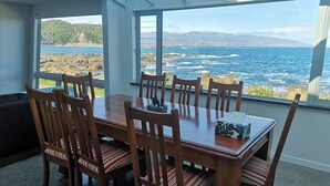 A dining area with a wooden table and chairs, large windows with a view of the sea and hills. - Esplanade Azure Horizon (Wellington)