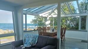 A modern living room with a large window overlooking the sea, a comfortable sofa, and a dining area with wooden furniture. - Esplanade Azure Horizon (Wellington)