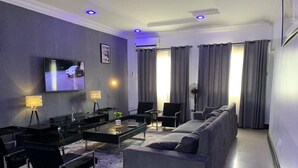 A modern living room with a large flat-screen TV, a sofa, a coffee table, and a dining area with chairs and a lamp. - 3 Bed Apartment at a Central Location (Ibadan)