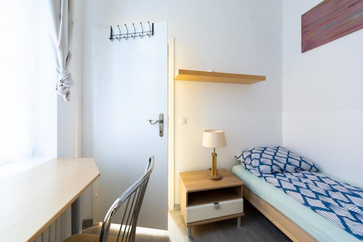 A compact room with a bed, a desk, a chair, and a bedside table with a lamp.