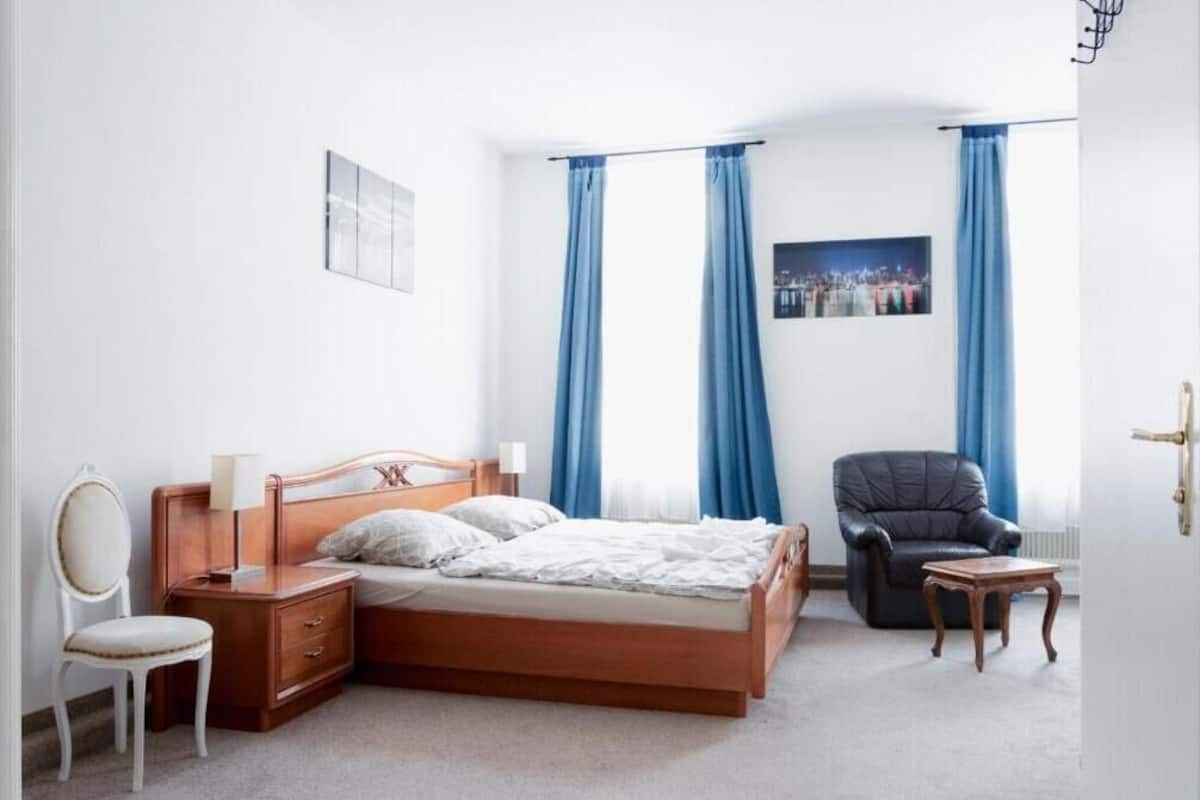 A bedroom with a wooden bed, a nightstand, a chair, a small table, and a cityscape painting on the wall.