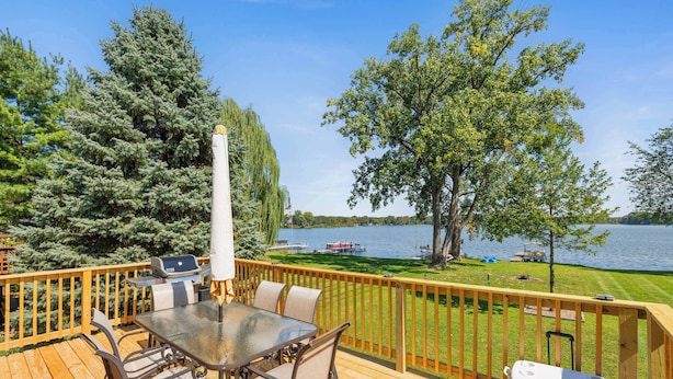 Eine Holzterrasse mit einem Glastisch und Stühlen mit Blick auf einen See, auf dem Boote und Bäume zu sehen sind.