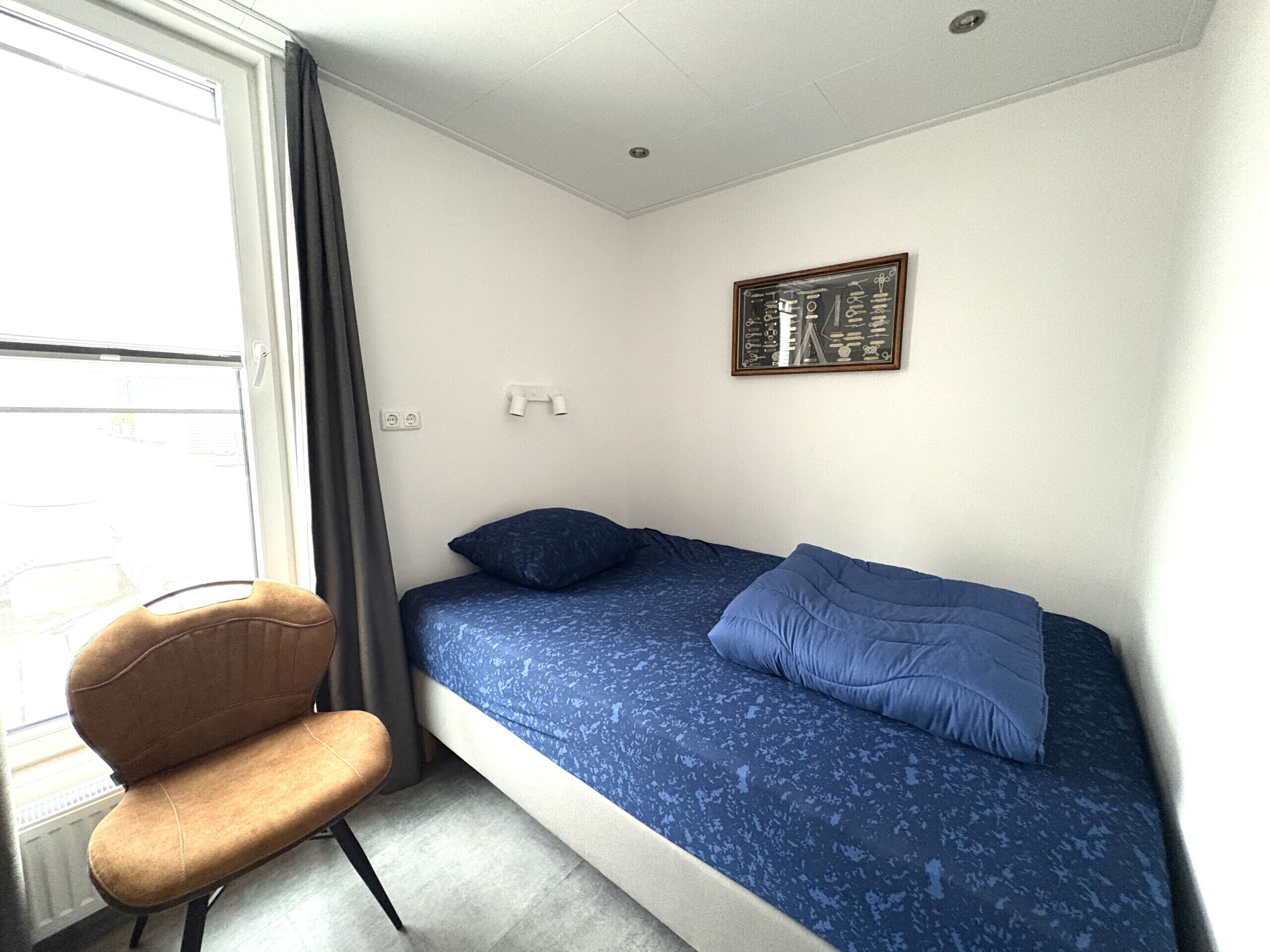 A small, single-bed room with a window, a chair, and a wall-mounted picture.