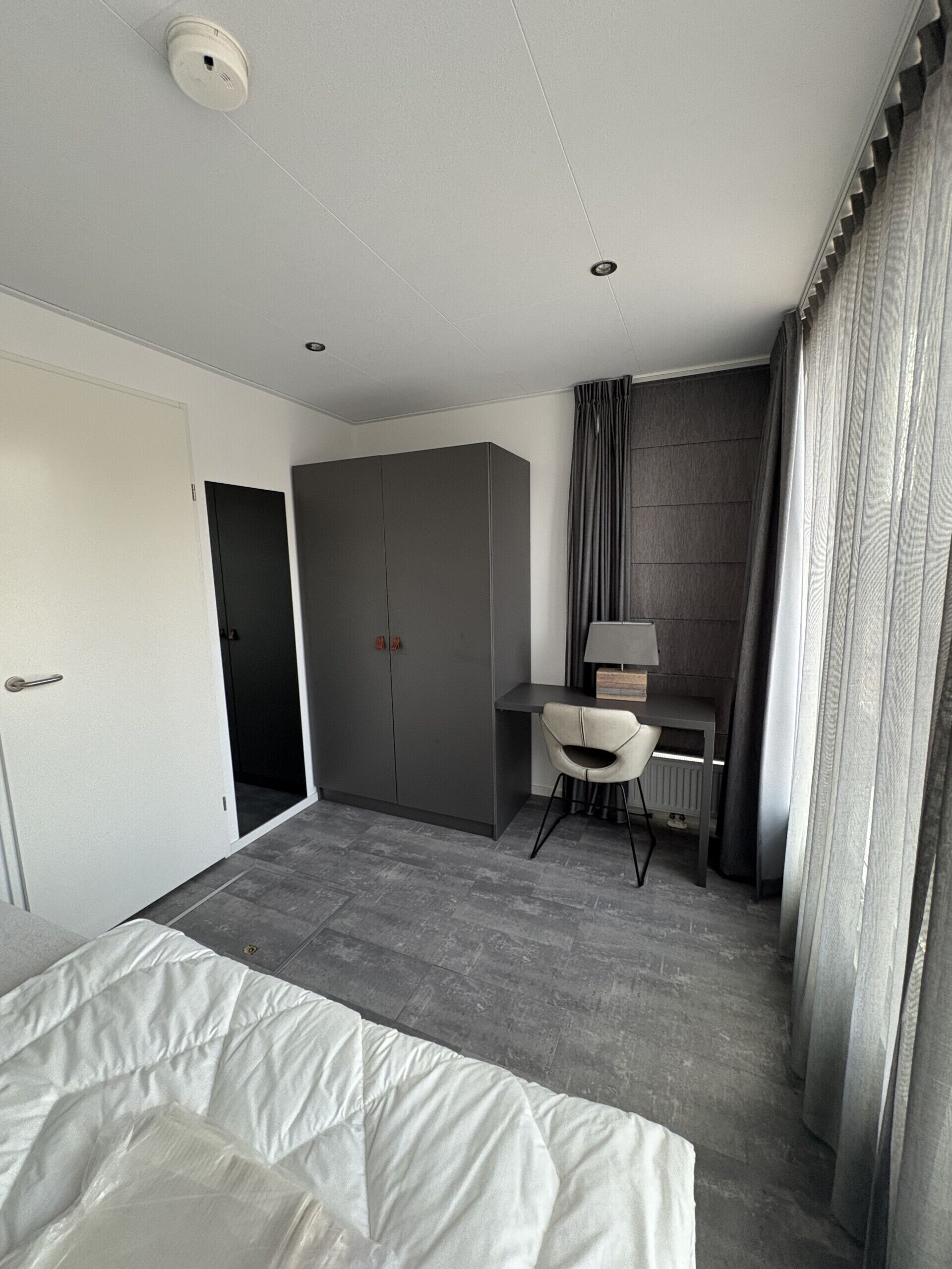 A modern hotel room with a bed, a desk with a chair, a wardrobe, and a bathroom door.