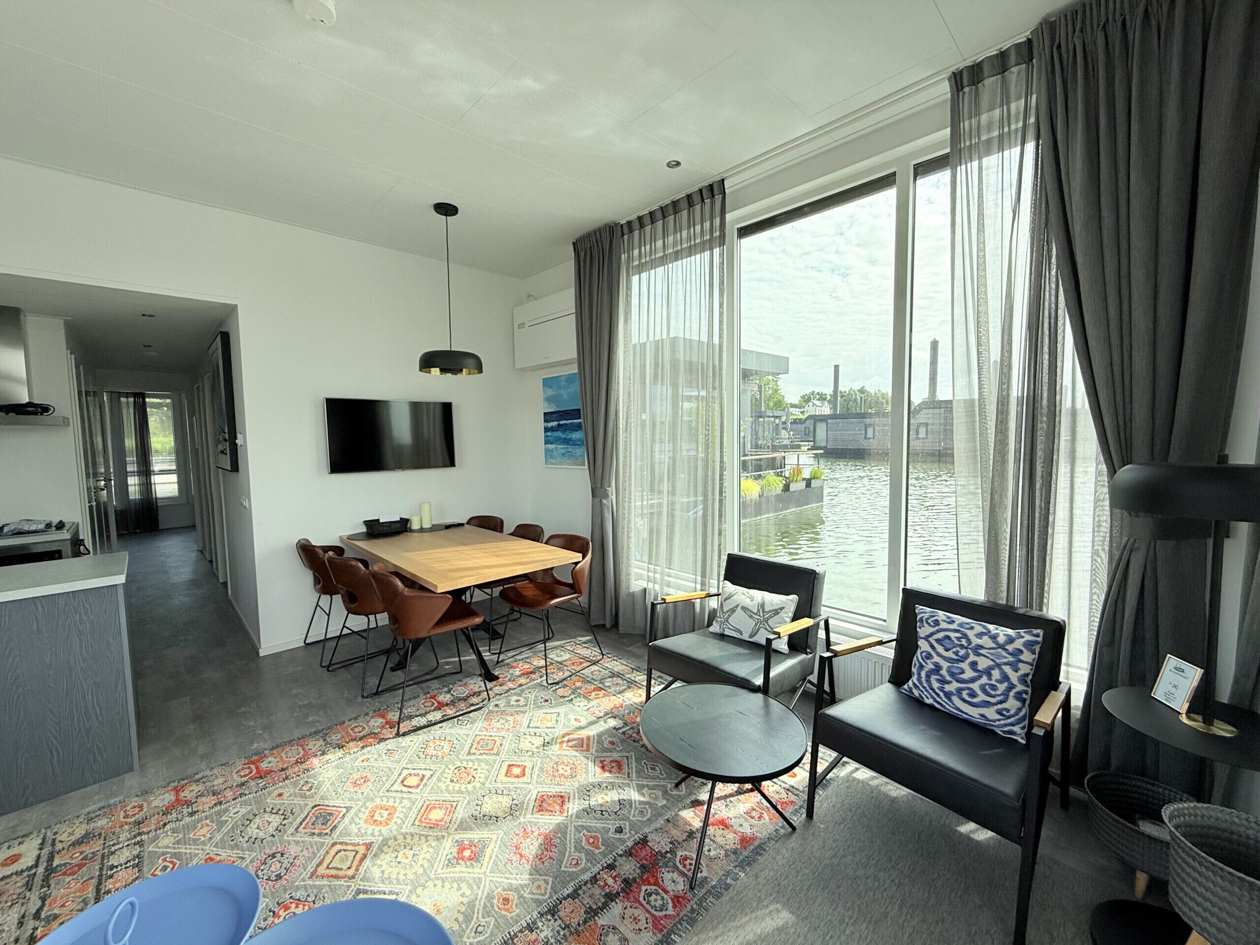A modern living room with a wooden dining table, comfortable seating, a flat-screen TV, and large windows offering a view of a water body and buildings.