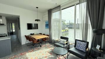 A modern living room with a wooden dining table, comfortable seating, a flat-screen TV, and large windows offering a view of a water body and buildings.