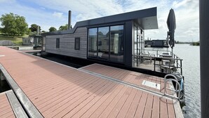 Ett modernt husbåt förtöjt vid en brygga, med ett trädäck, stora fönster och en täckt uteplats.