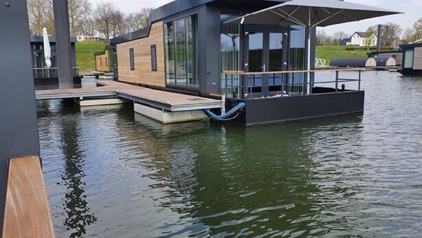 A modern floating house with a large glass facade, a wooden deck, and a covered outdoor area. - Maasterp - Watervillage 14 (Ohé en Laak)
