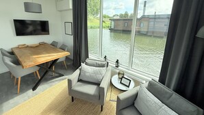 Una sala de estar moderna con una mesa de comedor de madera, sofás grises, un televisor de pantalla plana y un amplio ventanal con vistas a un cuerpo de agua.