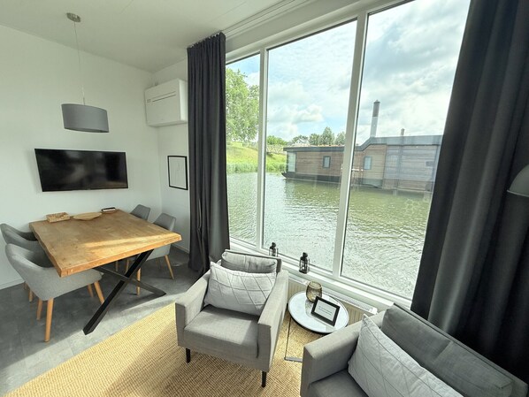 Een moderne woonkamer met een houten eettafel, grijze banken, een flatscreen-televisie en een groot raam met uitzicht op een waterpartij.