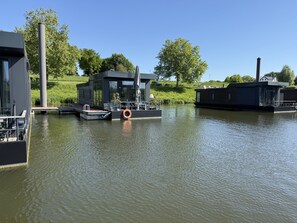 A modern houseboat docked at a canal with a grassy bank and trees in the background.