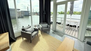 Una sala de estar moderna con grandes ventanales que dan a una masa de agua, amoblada con un sofá, sillones y una mesa de madera.