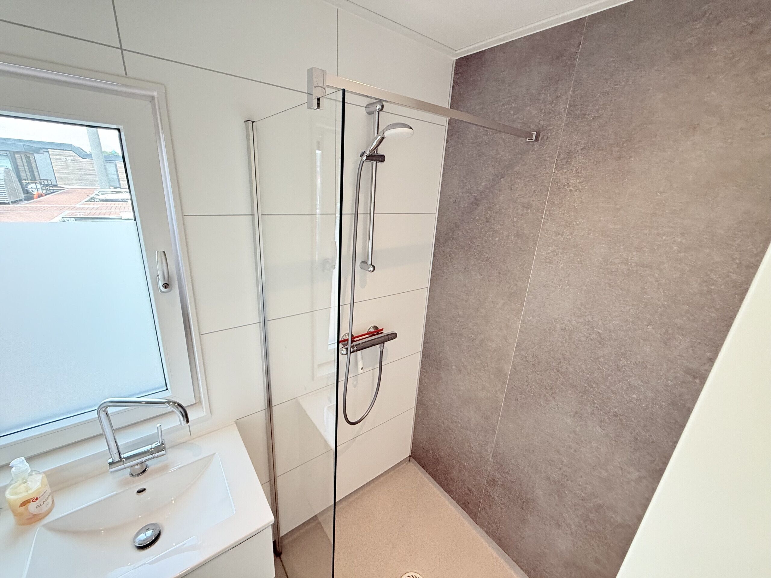 A modern bathroom with a glass shower enclosure, a white sink with a soap dispenser, and a view of the outside through a window.