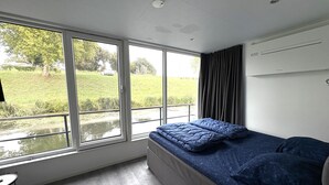 Un dormitorio moderno con un ventanal grande, una cama con ropa de cama azul, una mesita y vistas a un paisaje verde.