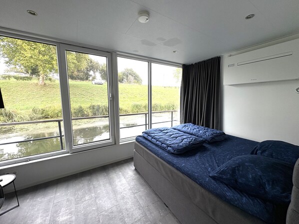 Une chambre moderne avec une grande fenêtre, un lit avec une literie bleue, une petite table et une vue sur un paysage verdoyant.