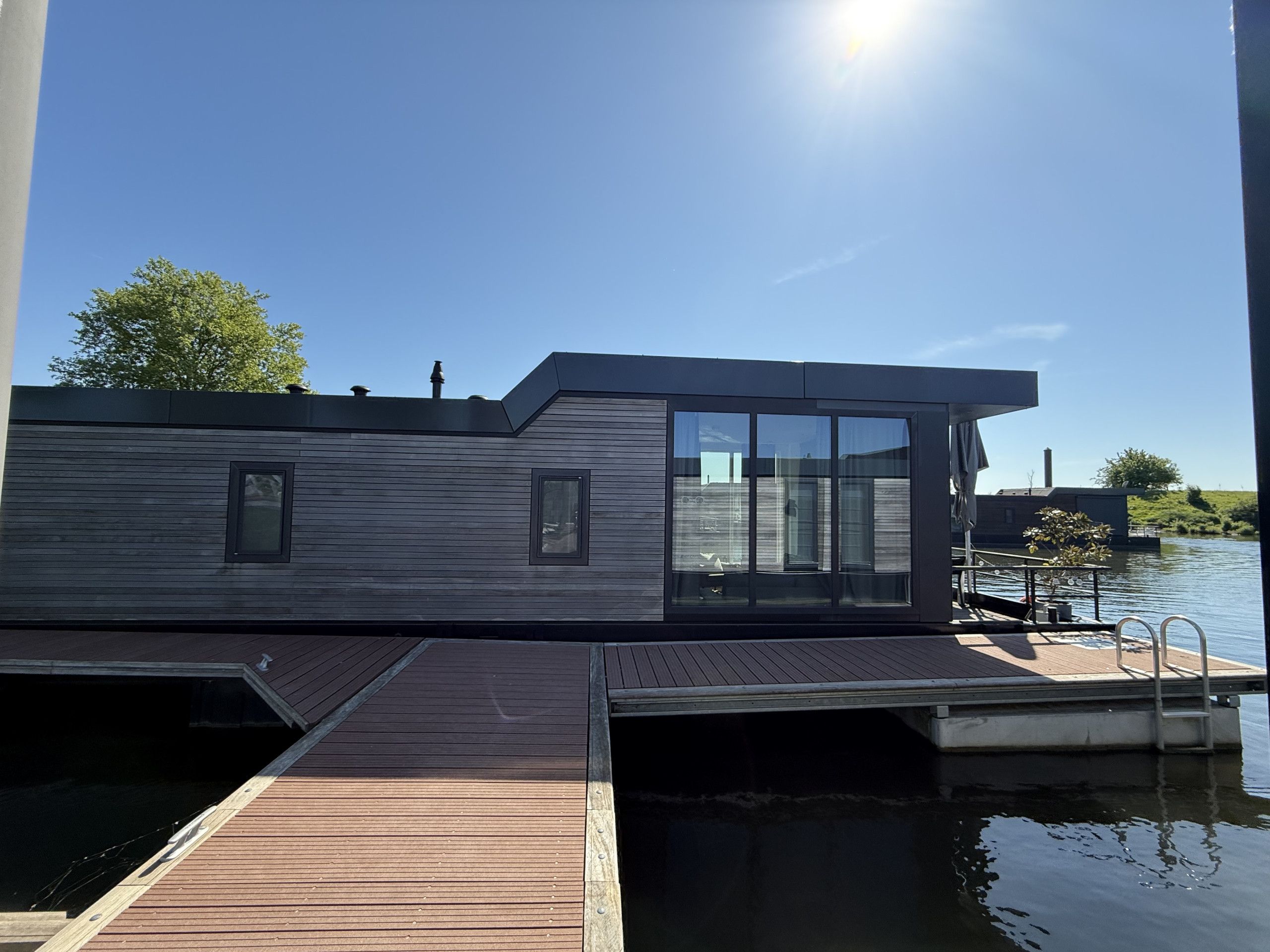 Una moderna casa flotante con terraza de madera, ventanas amplias y un muelle que se adentra en una masa de agua tranquila.