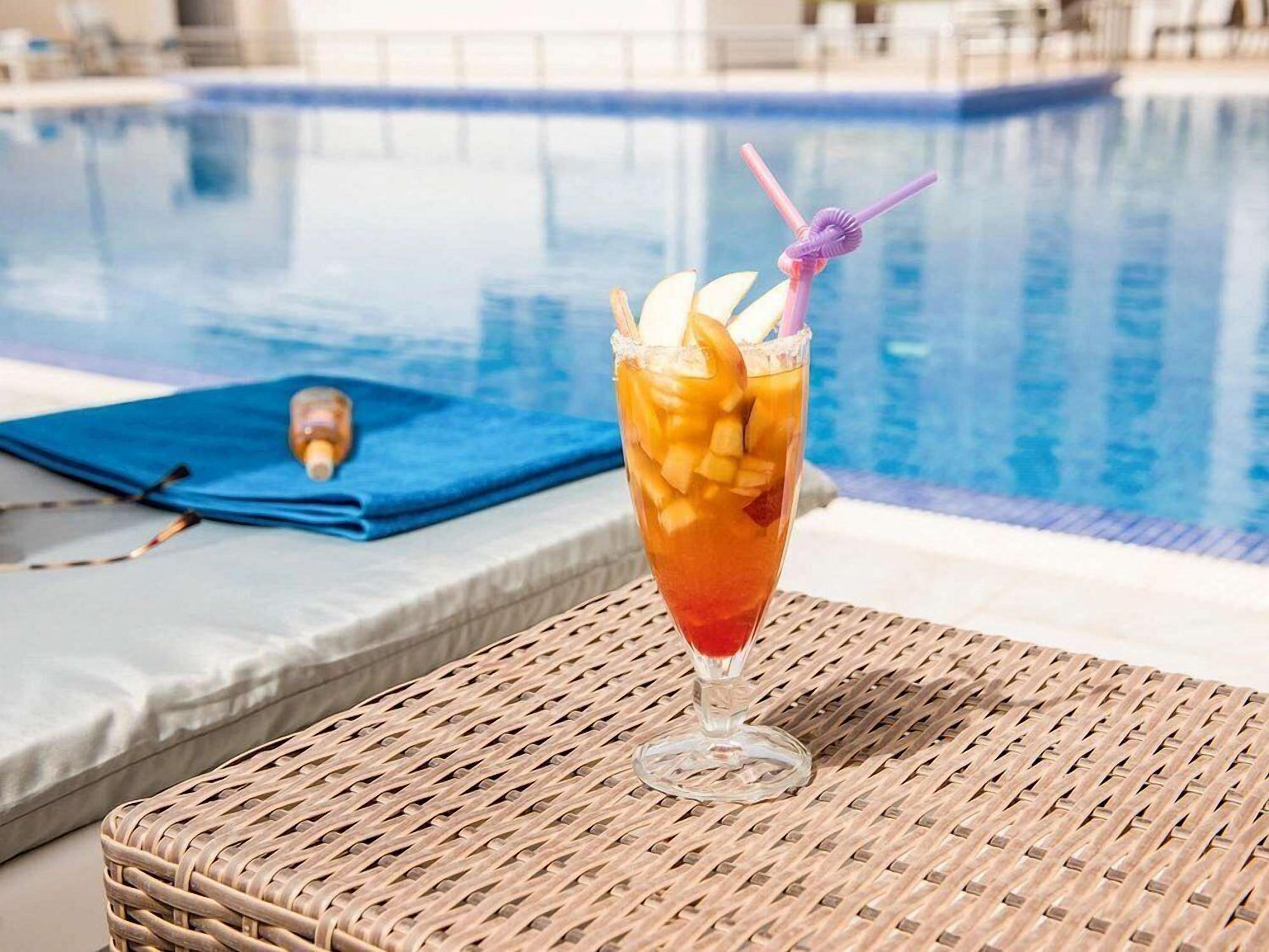 A cocktail with fruit garnish and a straw, placed on a wicker table by a poolside.