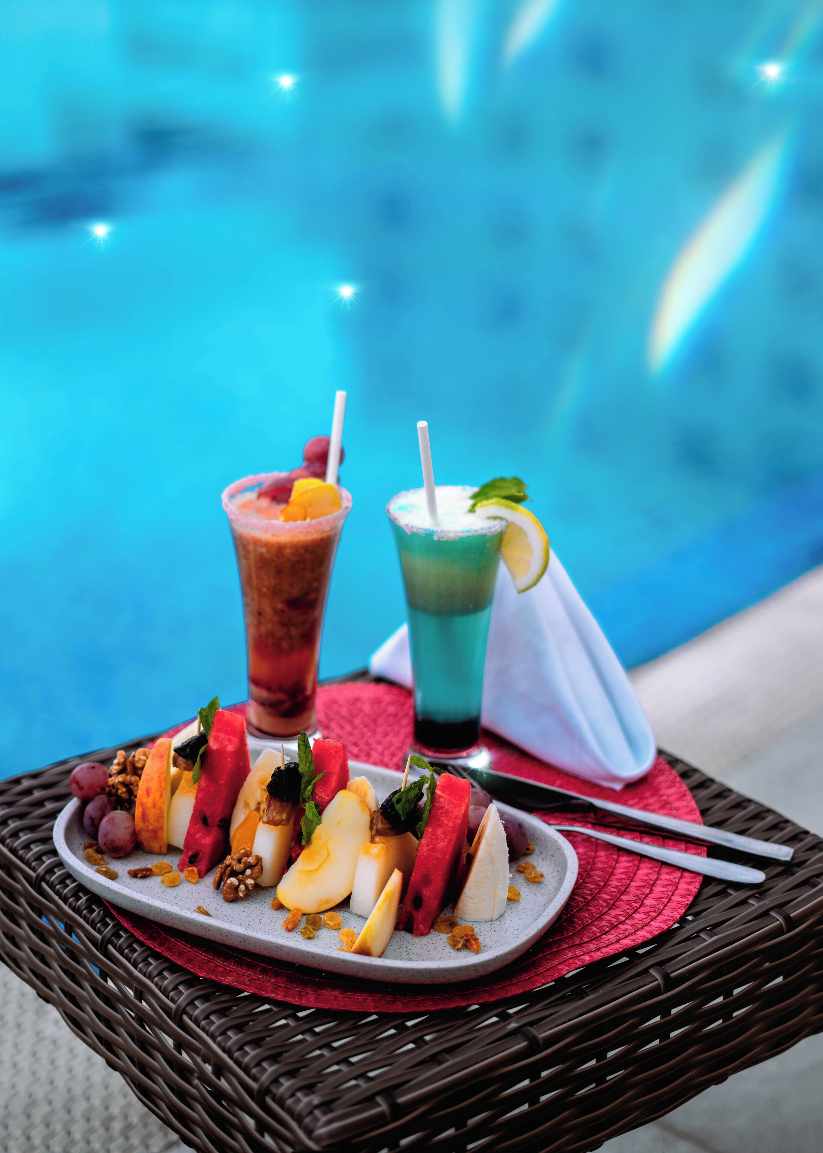 Una mesa con un plato de fruta y dos bebidas, una piscina al fondo.