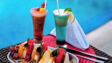 Una mesa con un plato de fruta y dos bebidas, una piscina al fondo.