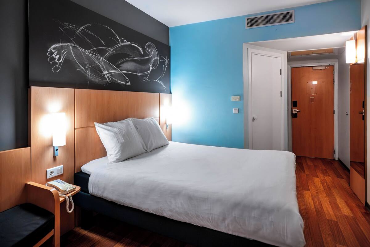A hotel room with a bed, bedside table with a phone, a wall-mounted light, and a door leading to another room.