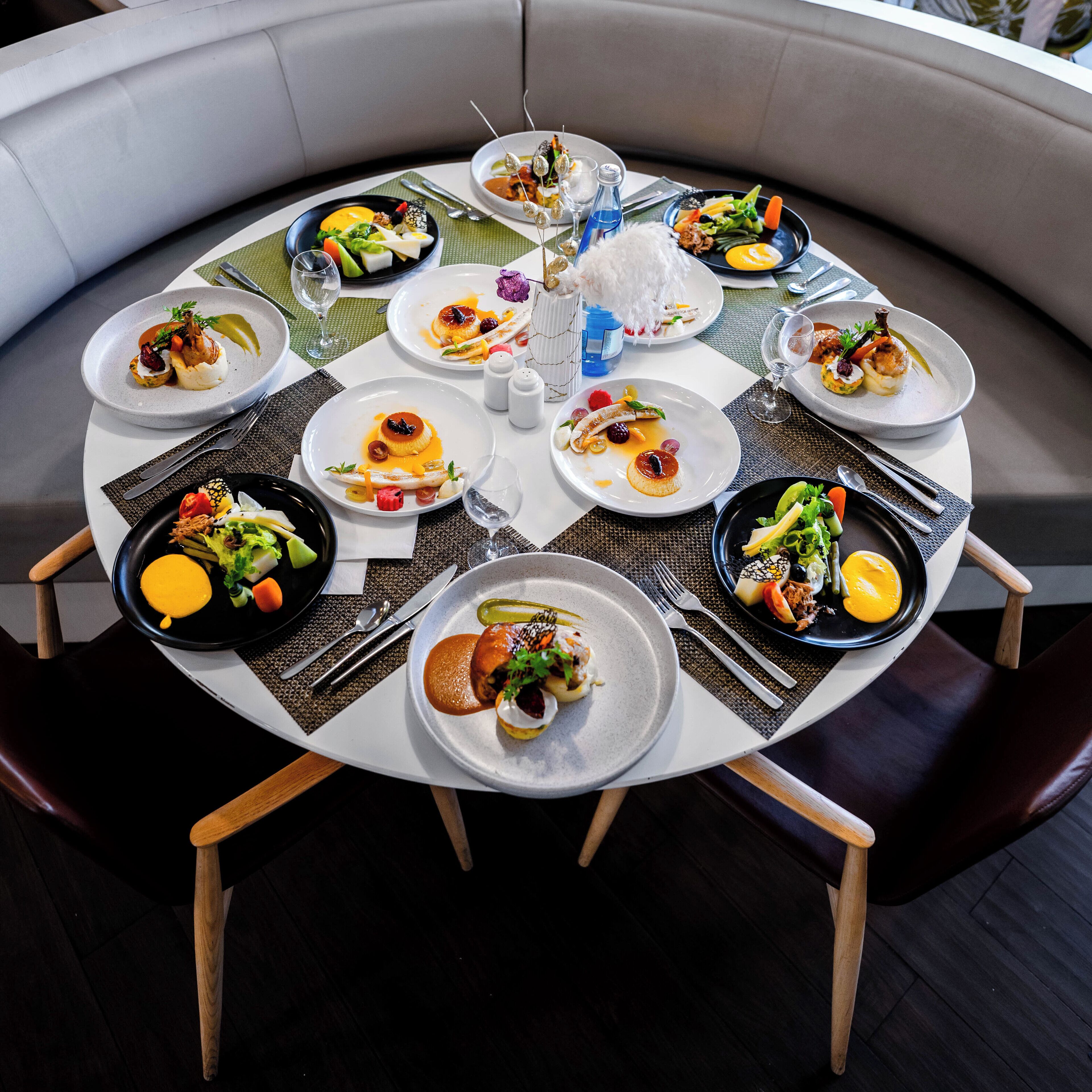 A round dining table with multiple plates of food, set with cutlery, glasses, and a bottle of water.