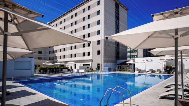A swimming pool with sun umbrellas, lounge chairs, and a modern building in the background. - Noxi Hotel (Setif)