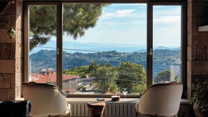 A room with a large window offering a view of a mountainous landscape. There are two chairs and a small table with a drink and a book. - Mila House (Ayvacik)