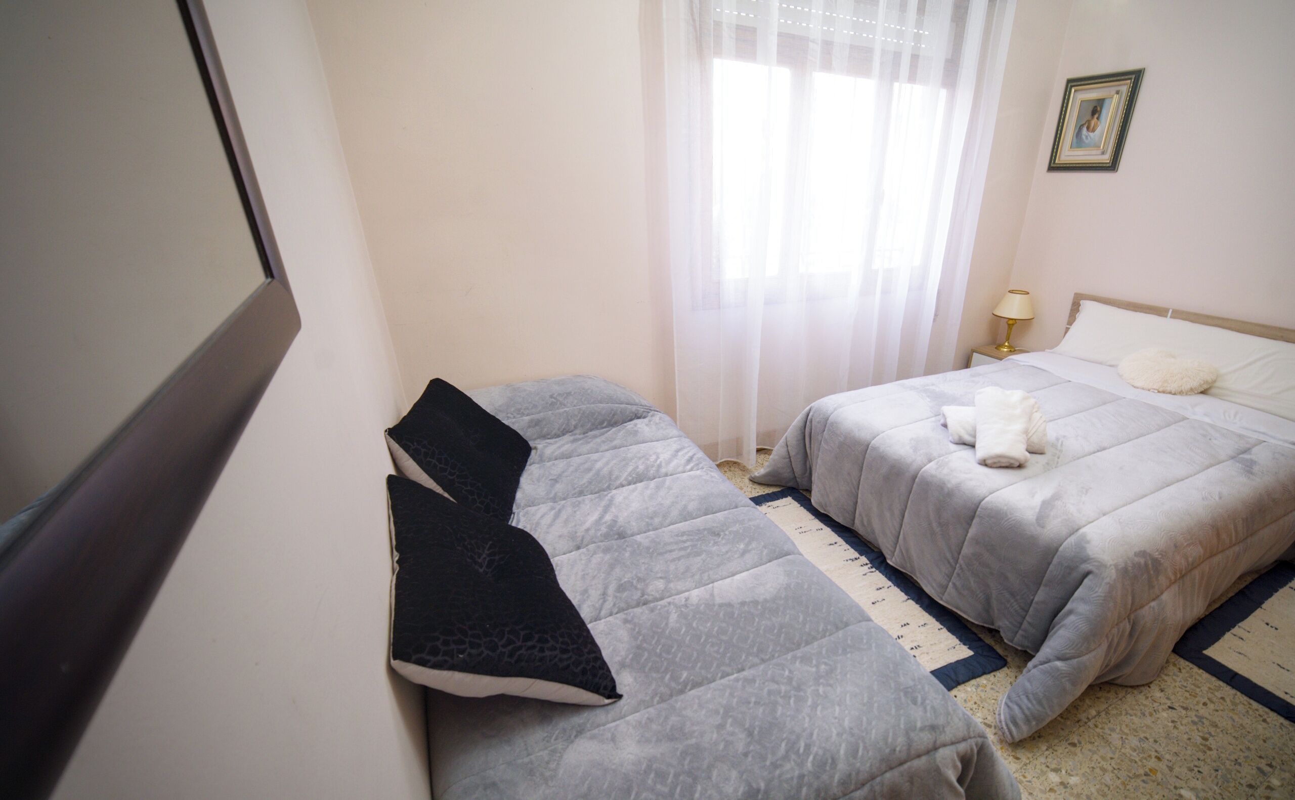 Une chambre d’hôtel avec deux lits, une table de chevet avec une lampe, un tableau encadré au mur et une fenêtre avec des rideaux légers.