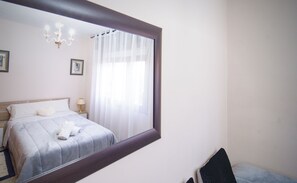 Une chambre avec un grand miroir, un lit recouvert de draps blancs, une table de chevet avec une lampe et une fenêtre ornée de rideaux légers.