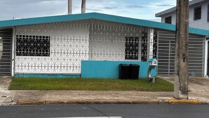 A building with a blue roof and white walls, featuring a metal security gate and a trash bin.