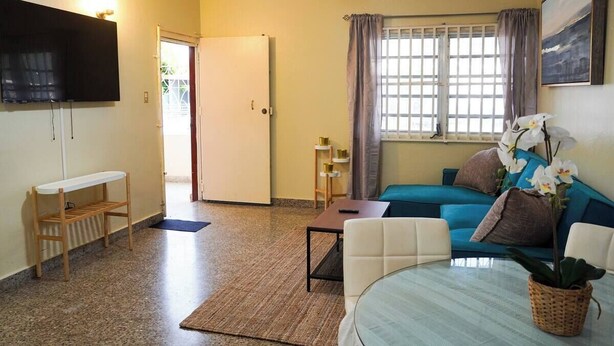 A living room with a sofa, a coffee table, a TV mounted on the wall, a window with curtains, and a door leading outside.