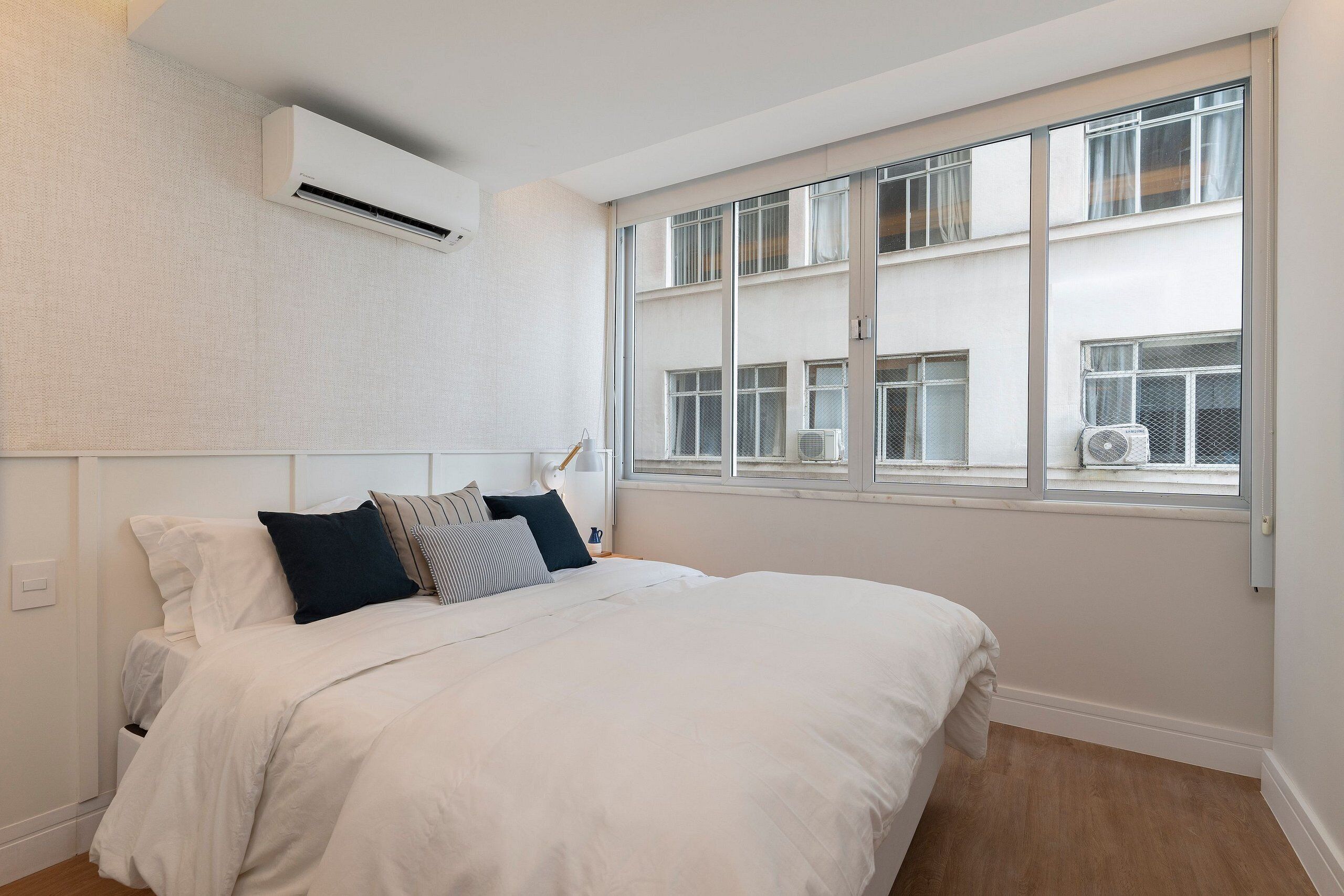 A bedroom with a large bed, white bedding, a window with multiple panes, and an air conditioning unit mounted on the wall.
