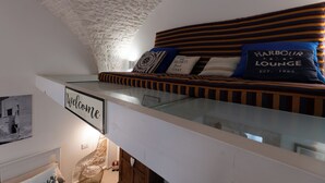 Une chambre de style loft dotée d’un lit surélevé avec une plateforme en verre, d’une pancarte « Bienvenue » et d’un coussin orné du mot « Harbour Lounge ».