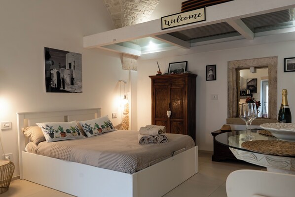 A bedroom with a bed, a wooden wardrobe, a glass table with a wine glass, and a 'Welcome' sign above the bed.