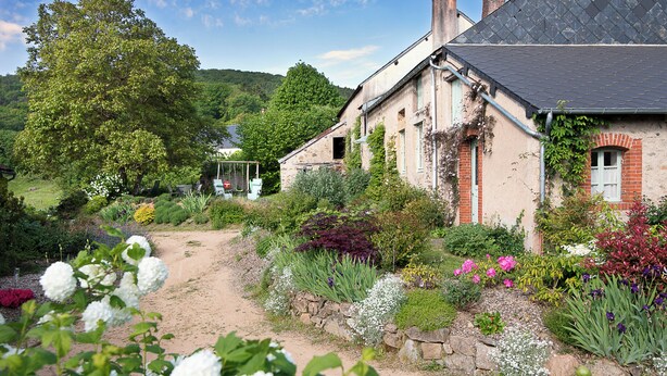 Ein rustikales Haus mit einem Feldweg, der davorhin führt, umgeben von üppiger Vegetation und blühenden Pflanzen.