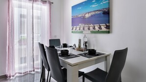 Een eetruimte met een tafel voor vier personen, stoelen, een schilderij van een kustlandschap en lichte gordijnen.