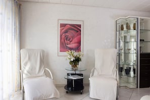 A room with two white chairs, a glass table with a vase of flowers, a framed picture of a rose, and a glass cabinet with shelves.