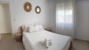 Una cama bien hecha con sábanas blancas y una toalla doblada, dos espejos en la pared, una mesita redonda y una ventana con cortinas de encaje.