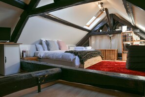 Un dormitorio tipo loft con techo inclinado, una cama con almohadas, una alfombra roja, una estantería y un escritorio con una lámpara.
