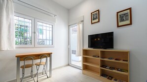 A room with a TV on a wooden stand, two foldable chairs, a bookshelf with books, and a window with curtains.