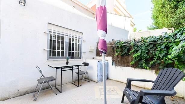 An outdoor patio area with a table and chairs, a black chair, a white wall, a window with bars, and a covered umbrella.