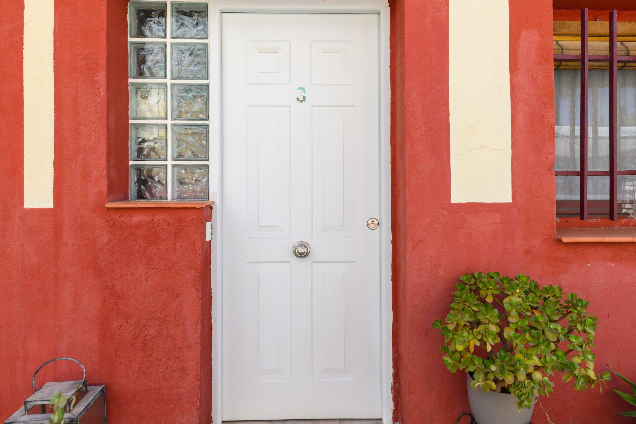 Une porte blanche portant le numéro « 3 », située sur un bâtiment rouge avec une fenêtre au-dessus et un pot de fleurs sur le côté droit.