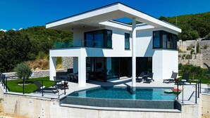 A modern two-story house with a large swimming pool, outdoor seating, and a view of the surrounding landscape.
