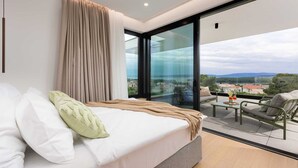 A modern bedroom with a large bed, a view of the city through floor-to-ceiling windows, and a balcony with outdoor seating.